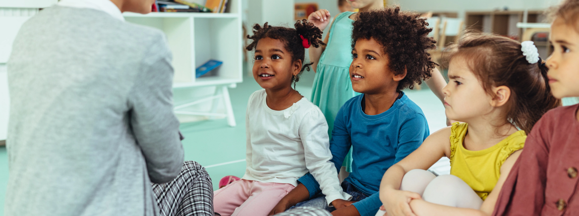 A daycare teacher is interacting with a diverse group of engaged, young students.