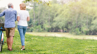 A senior couple walking through a park, with the male pushing a bike.