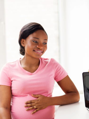 A smiling African American pregnant woman at a health clinic with a female doctor standing at a computer in the foreground.