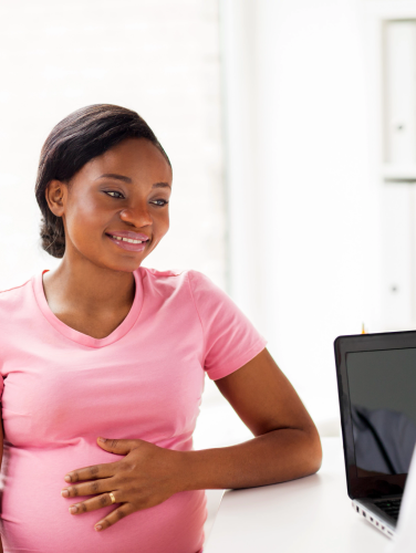 A smiling African American pregnant woman at a health clinic with a female doctor standing at a computer in the foreground.